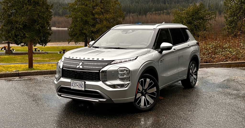 2026-Outlander-PHEV - Ingersoll Voice Front-three-quarter view of a silver Mitsubishi Outlander SUV parked on a wet pavement with trees and a lake in the background, daytime outdoor scene.