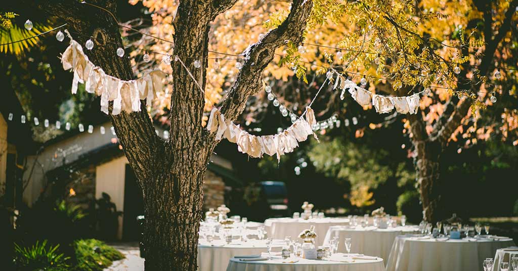 Dining-event-formal • Troy Media Outdoor garden event setup with round tables dressed in white linens and string lights between trees at dusk.
