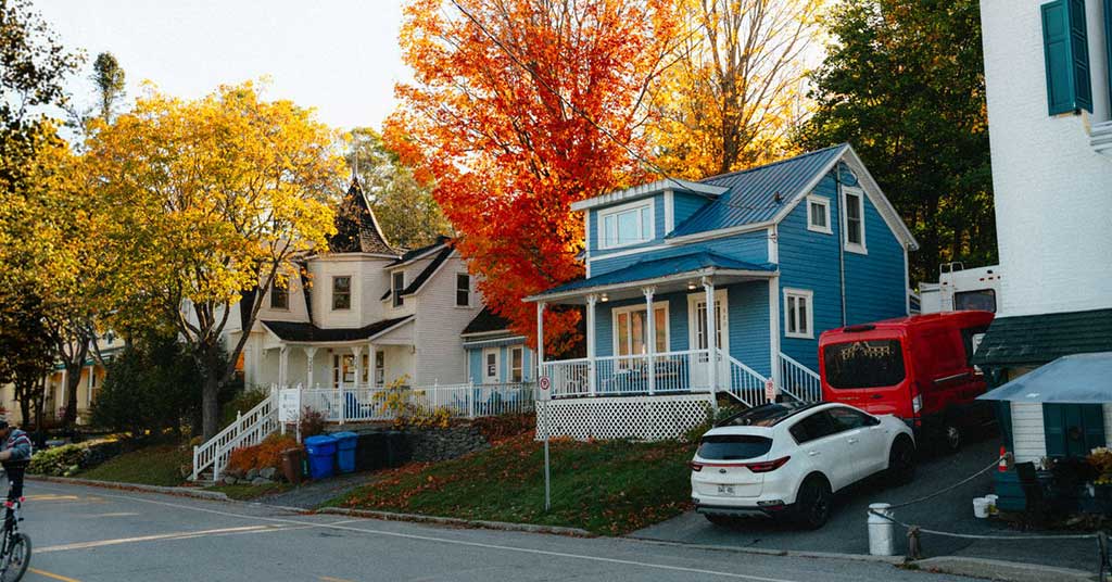 Housing-neighbourhood • Troy Media Blue two-story house with a white porch on a fall-colored street, orange and yellow trees, and a white car with a red truck nearby.
