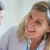 Woman (adult) with blonde hair smiles at a young girl with colorful hair clips, suggesting a friendly, caring interaction in a classroom or clinic setting.