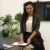 Professional woman sits on a conference table with a laptop and notebook in a bright office with plants nearby.