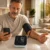Man in a casual gray shirt sits at a kitchen island, checking blood pressure with a digital monitor while looking at his smartphone, cuff wrapped around his left arm.