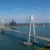 Aerial view of a white cable-stayed bridge spanning a wide river, with a city skyline in the distance on a sunny day.