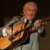 Older man in a light gray suit playing an acoustic guitar on stage, singing into a microphone.