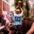 Crowd of fans on a street celebration; a footballer in a blue striped jersey (number 10) raises a golden trophy above his head while fans film and cheer.